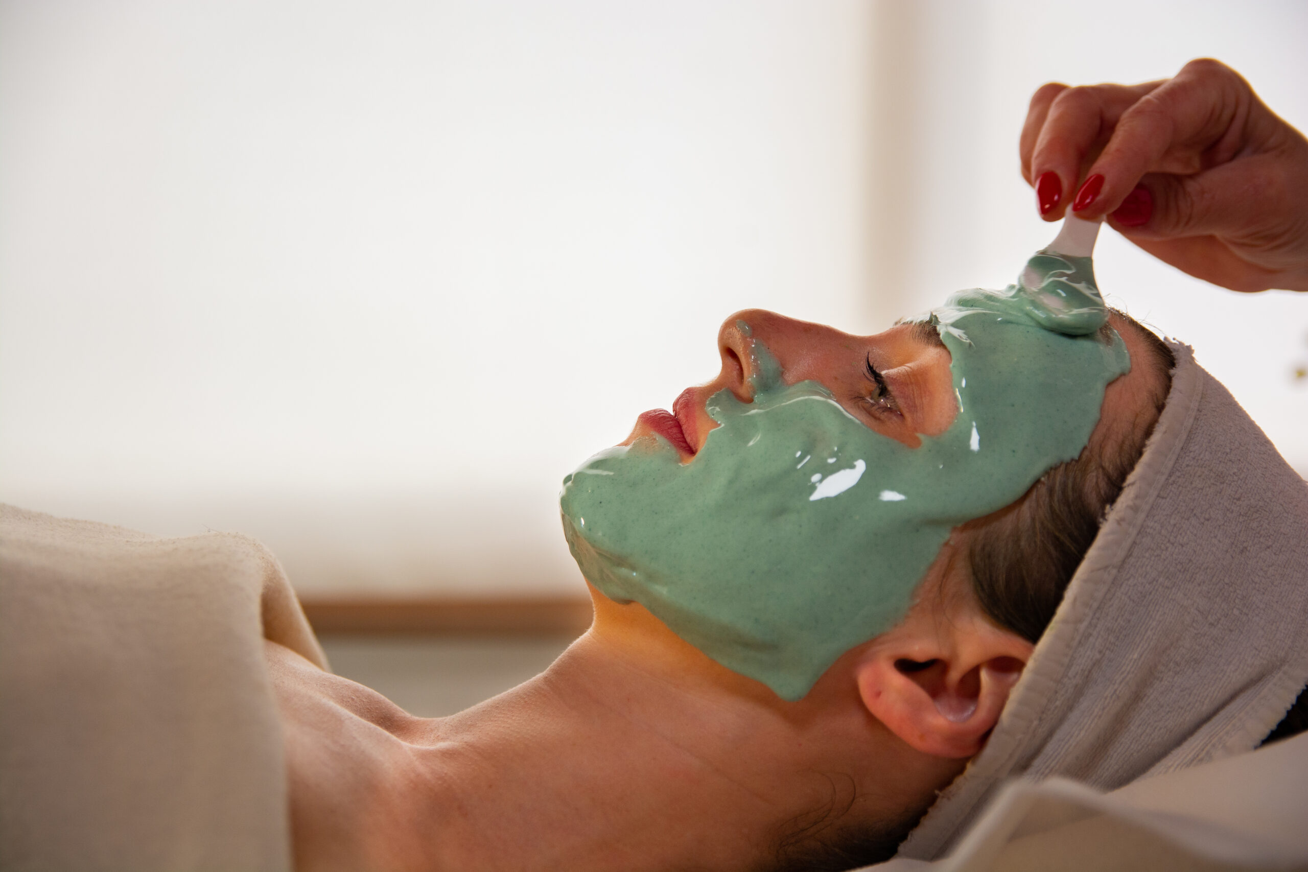 Close-up of young woman lying down and getting facial mask beauty treatment by cosmetician