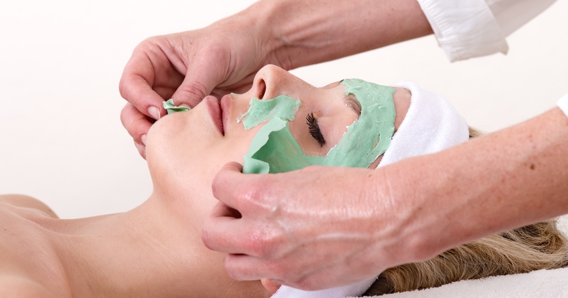 Beautician peeling off the green beauty facial mask on a laying and relaxed beautiful blond woman against a white background